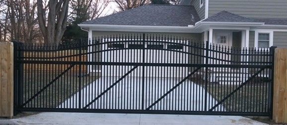 Black metal driveway gate with wooden posts, in front of a house.
