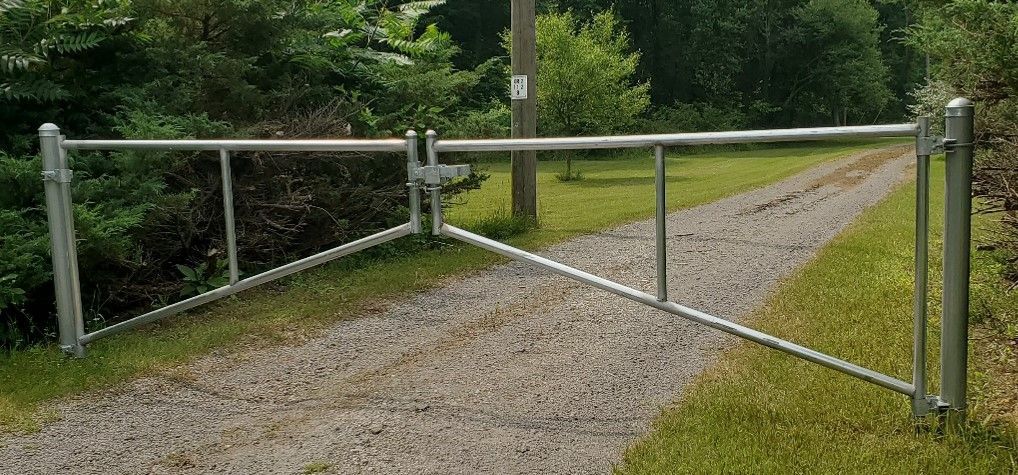 A silver metal gate across a gravel driveway in a grassy area with trees in the background.
