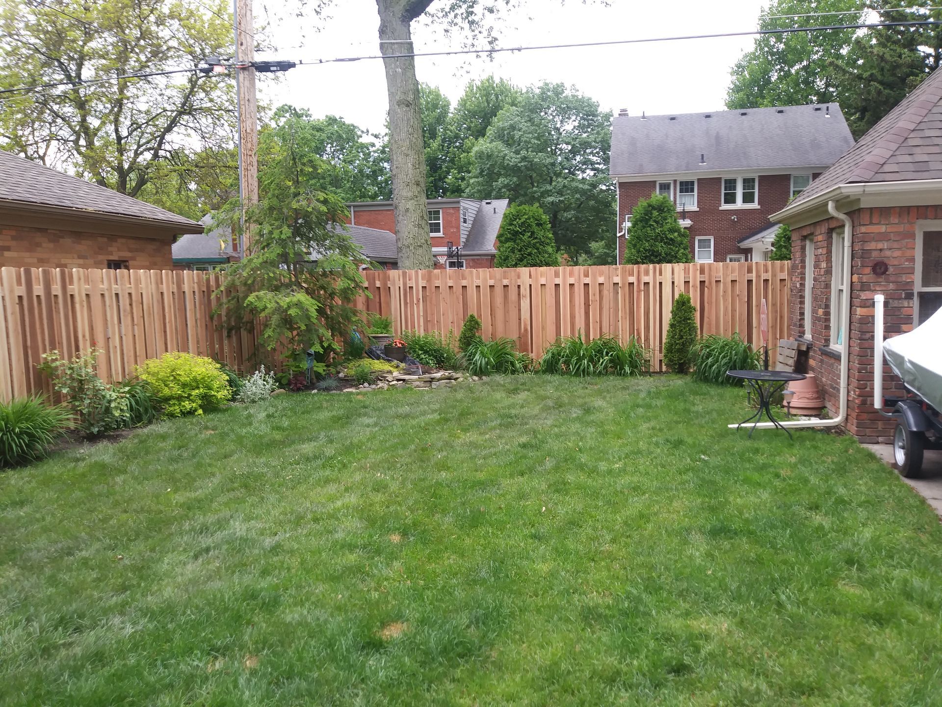 Lush green backyard with a wooden fence, trees, and a brick house on a cloudy day.