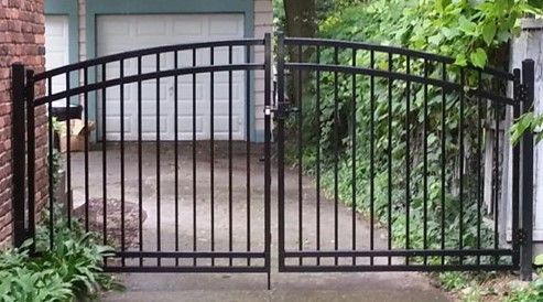 Black metal driveway gate in front of a garage, with arched top and vertical bars.