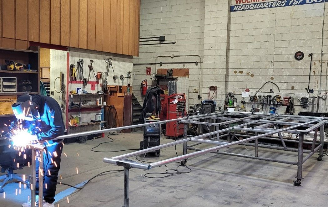 Welder in a shop, working on a metal frame with bright sparks. Tools and equipment in the background.