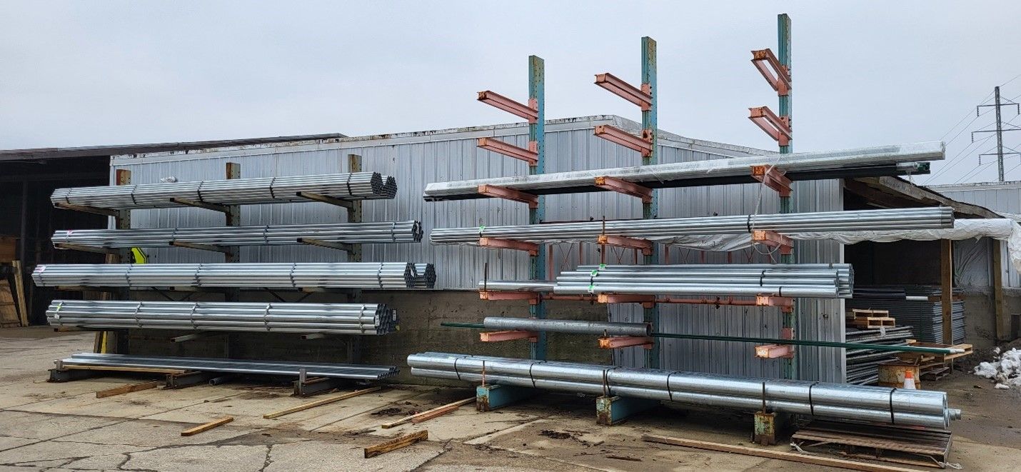 Metal pipes stored on shelving outside a building. Overcast, brown ground.