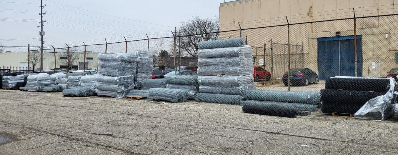 Piles of chain-link fencing rolls stored outdoors near a building with a fence.