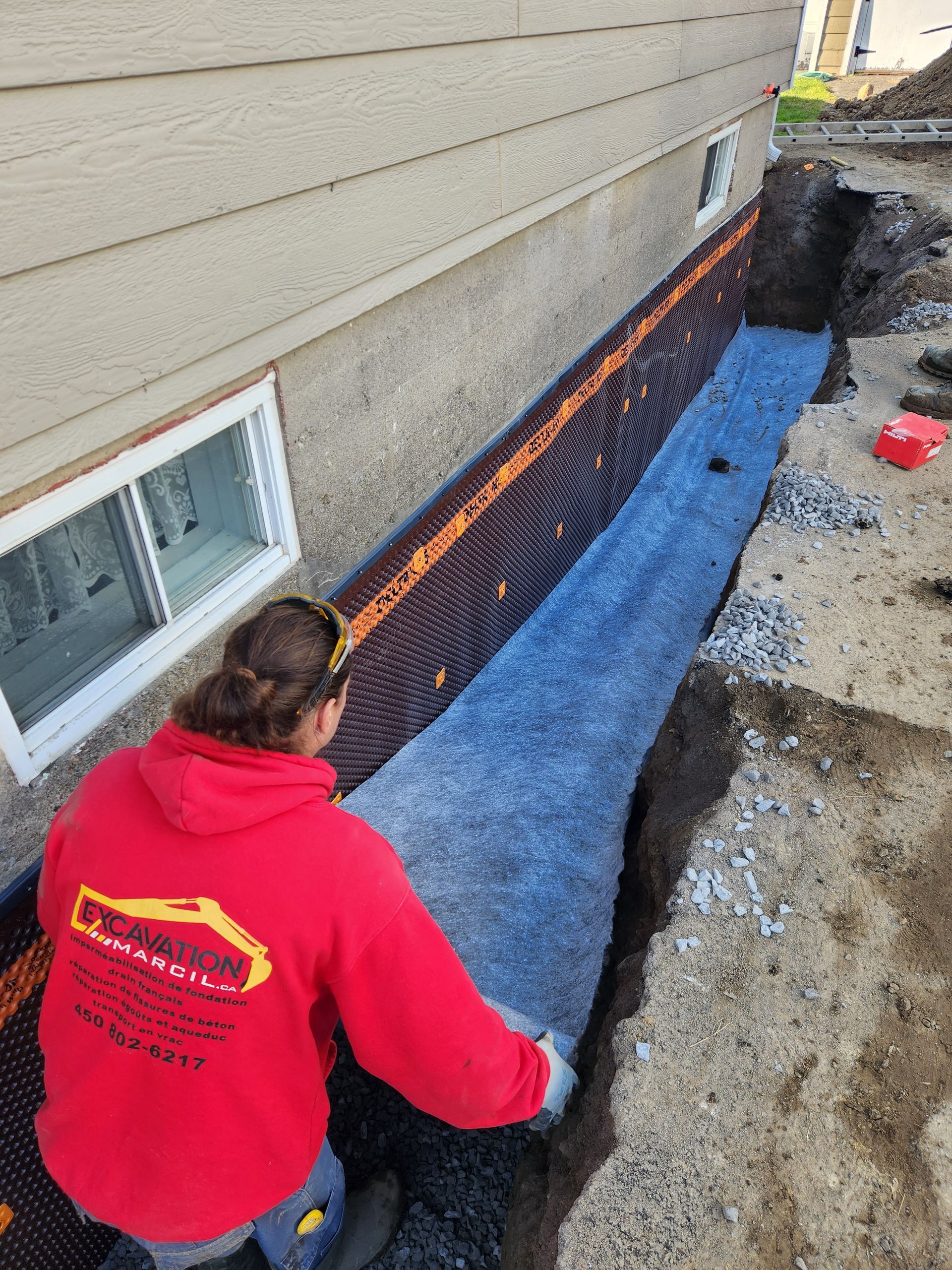 Une femme portant un sweat à capuche rouge travaille sur un système de drainage sur le côté d'une maison.