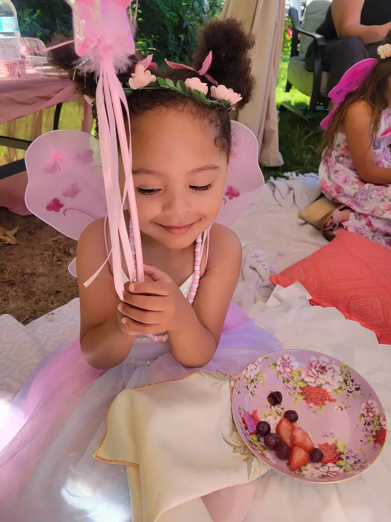 A little girl dressed as a fairy is sitting on a blanket with a bowl of fruit.