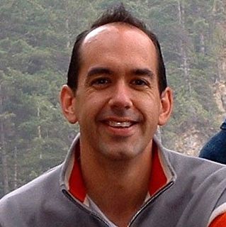 A man is smiling for the camera in front of a forest.