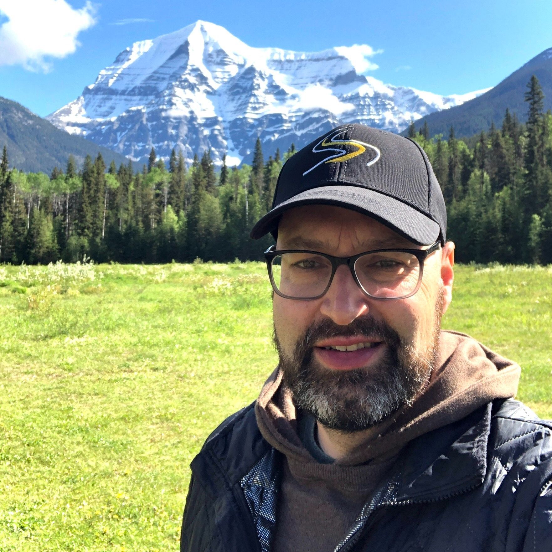 A man wearing glasses and a hat is standing in a field with a mountain in the background