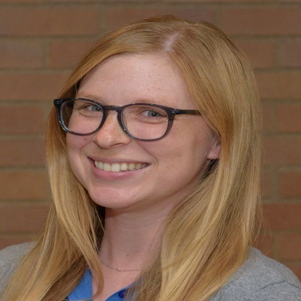 A woman wearing glasses is smiling in front of a brick wall.