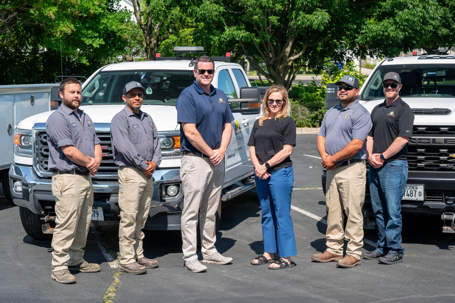 A group of seven people standing in front of two work trucks on a sunny day. They appear to be a work crew, smiling at the camera.
