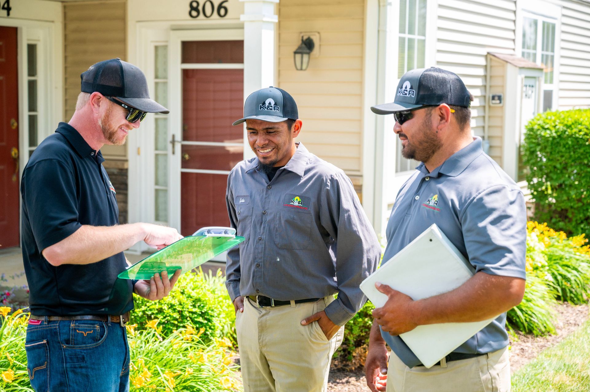 Three men in uniform, possibly landscaping professionals, reviewing a document outdoors in front of a house. One man holds the document, another smiles, and the third holds a clipboard.