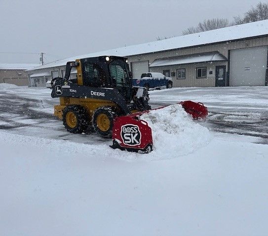 A yellow skid steer with a snow plow clears snow from a parking lot on a snowy day.