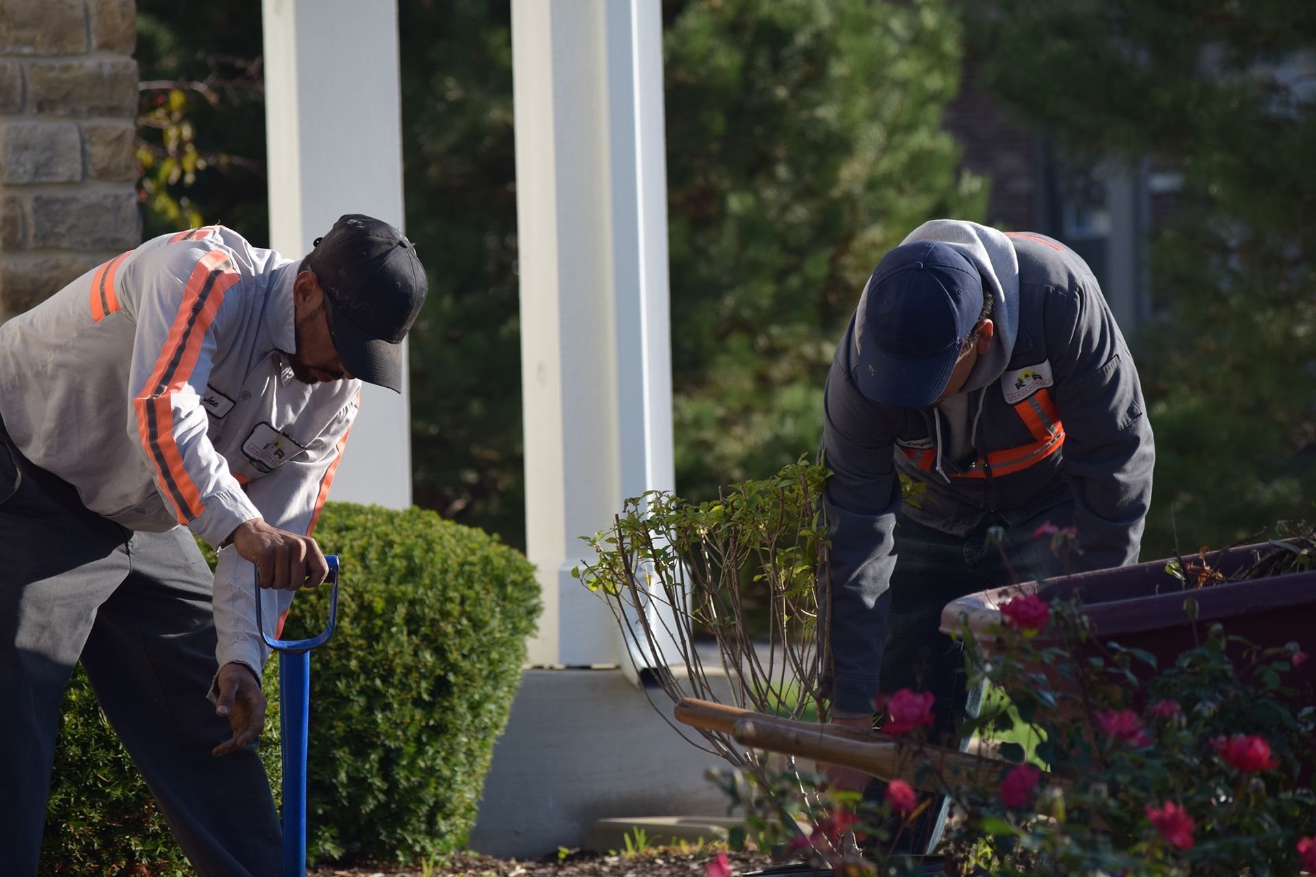 Two workers outside, one with a tool near a bush and another bending over a flower bed.
