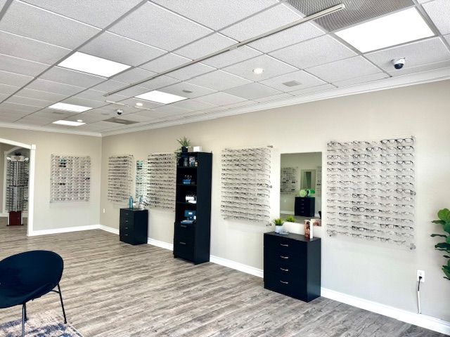 Optical shop interior, eyeglasses on display, neutral walls, wood-look flooring, black cabinets, overhead lighting.