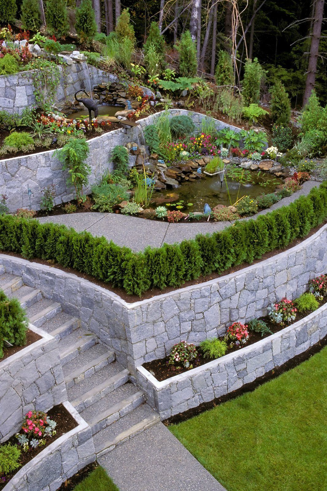 Stone terraced garden with paths, stairs, pond, and diverse colorful flowers and plants.