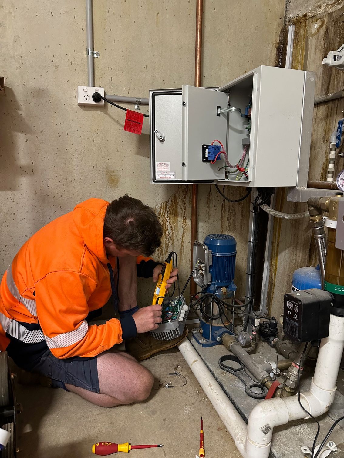 Technician in orange jacket kneels by a boiler, inspecting exposed pipes and electrical controls  — PUMPtek in Bendigo, VIC