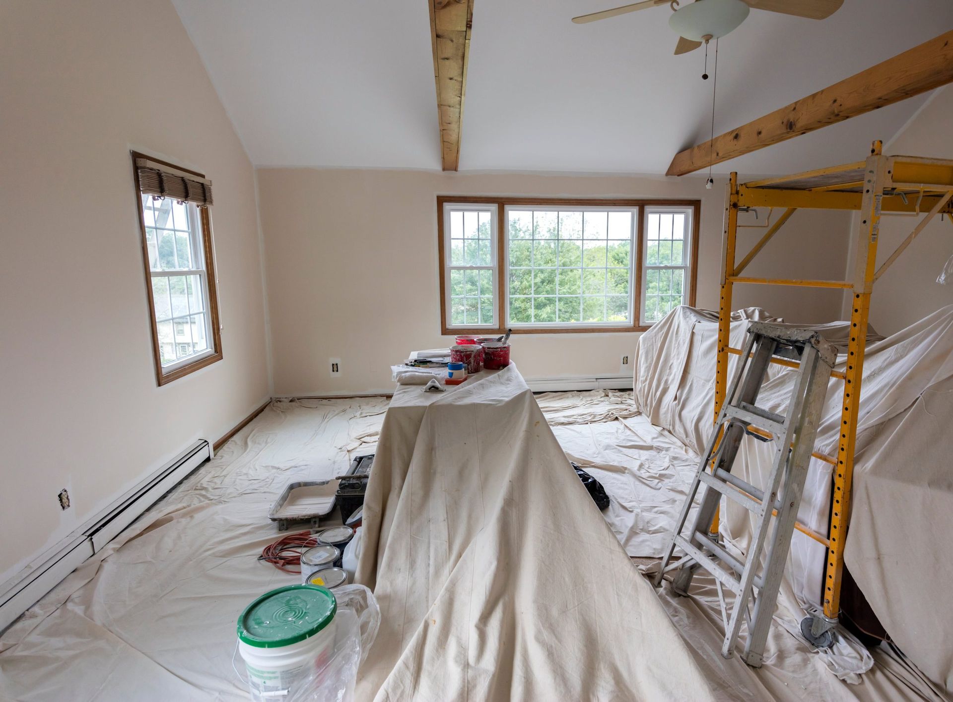 A room under renovation with beige walls, exposed wooden ceiling beams, a yellow scaffolding tower, and drop cloths.