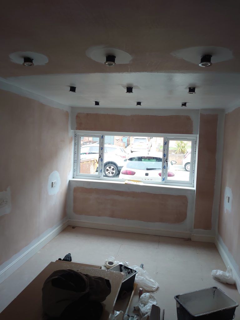 An interior room under renovation with fresh plaster on walls and ceiling, featuring recessed lighting and a bay window.