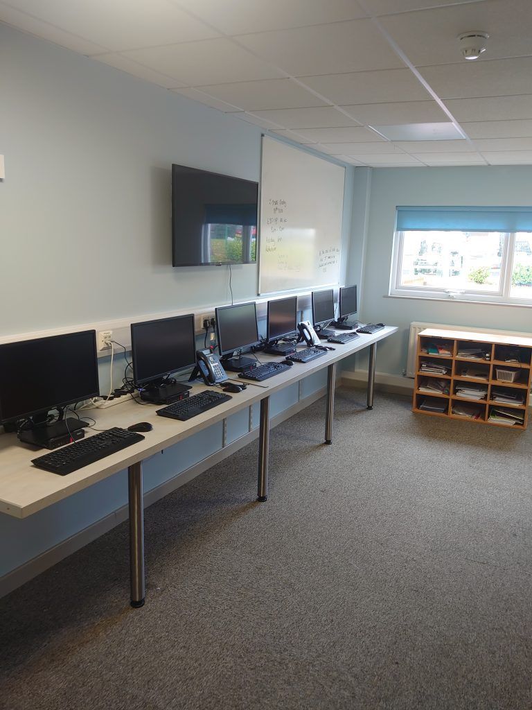 A row of computer workstations arranged along a pale blue wall in a classroom or office setting.