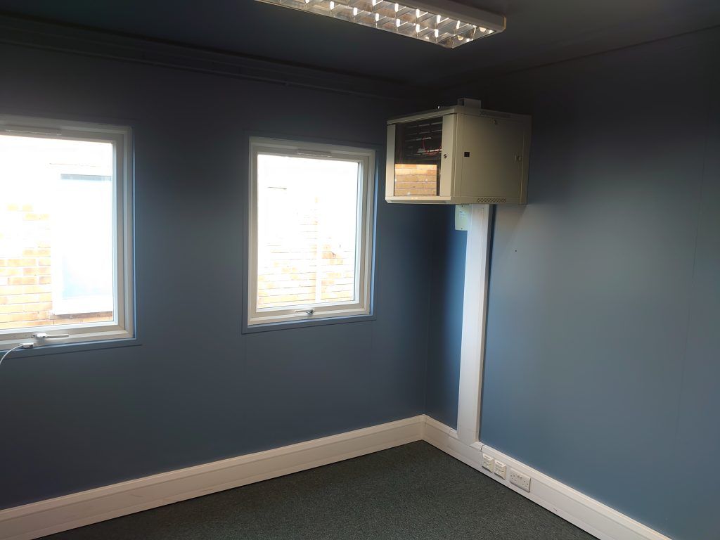 Empty room with dark blue walls, two windows, and a wall-mounted server cabinet with white casing on the right wall.