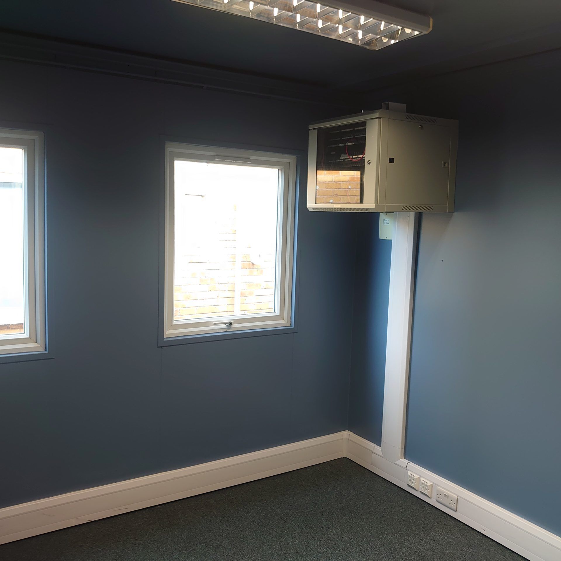 A small, empty office with blue walls, two windows, grey carpet, and a wall-mounted server rack.