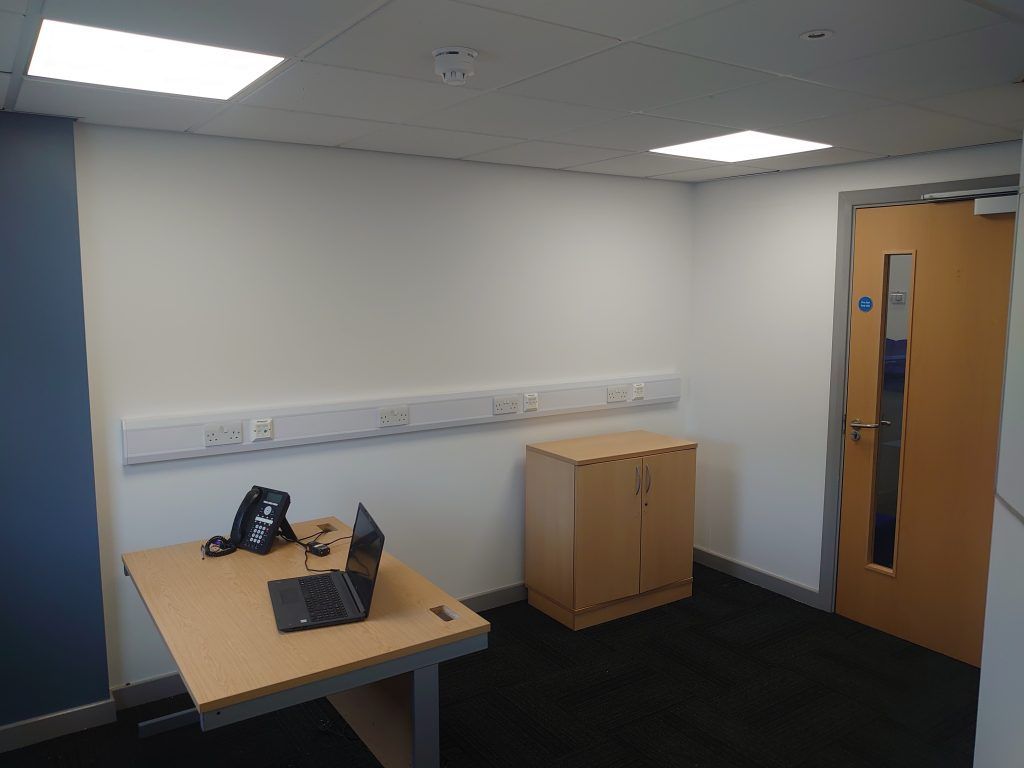 An office room with a desk, laptop, phone, a light wood cabinet, and a door against a white wall.