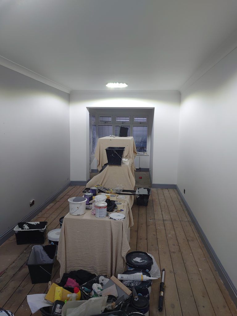An interior room under renovation with freshly painted white walls, bare wood floors, and drop cloths covering furniture.