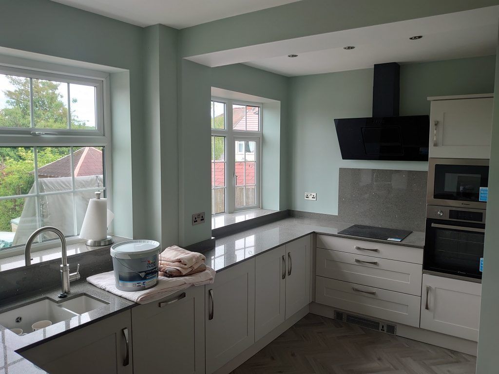A bright, modern kitchen with sage green walls, light cabinetry, stone countertops, and a black stove hood.