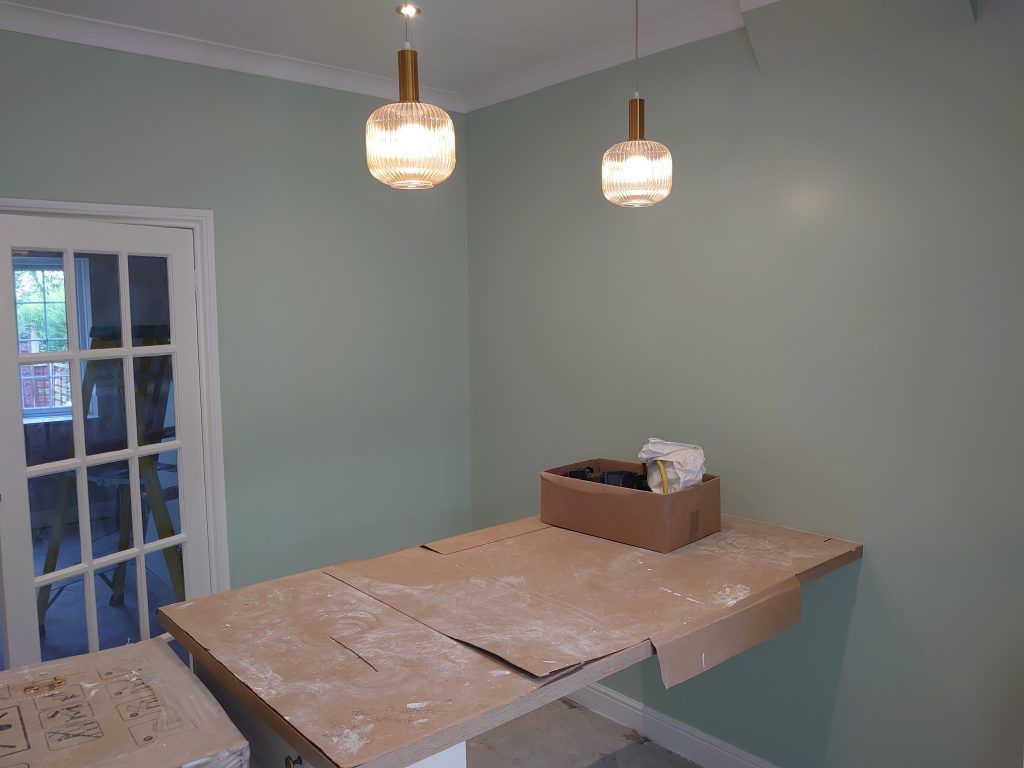 A room with pale green walls, two hanging pendant lights, and a wooden table covered in brown paper and supplies.