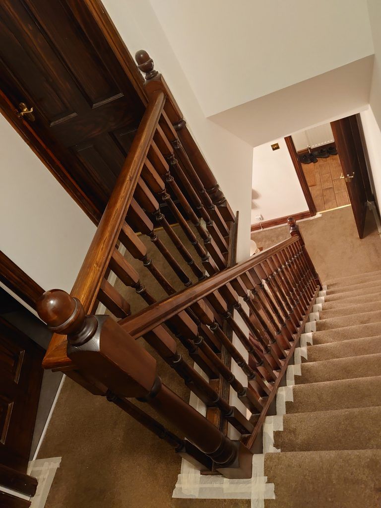 A high-angle view of a wooden staircase with dark railings, beige carpet, and white walls, leading to a landing.