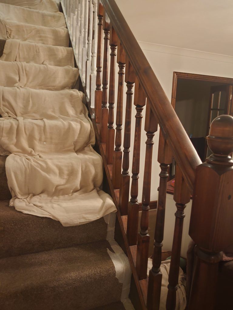 A staircase with brown carpet, partially covered by white drop cloths, and a wooden banister with vertical spindles.