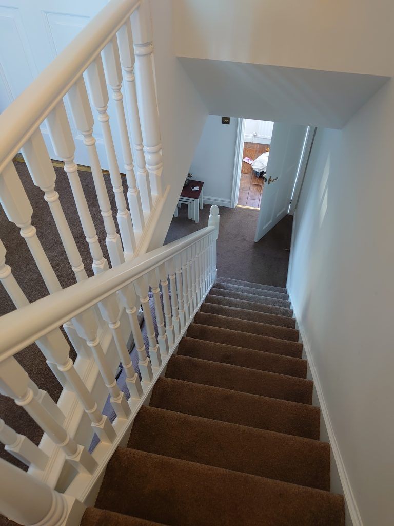 A high-angle view looking down a carpeted staircase with white railings, leading to a doorway opening into a lit room.