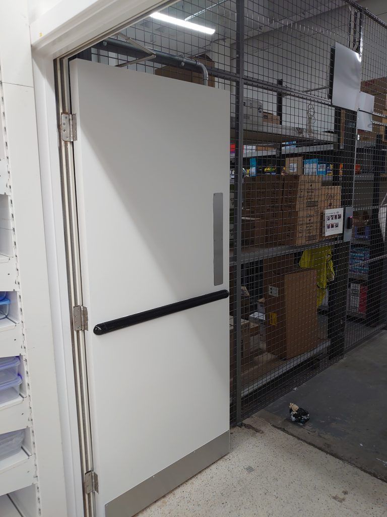 A white commercial door with a black push bar and silver kick plate, set in a frame within a warehouse environment.