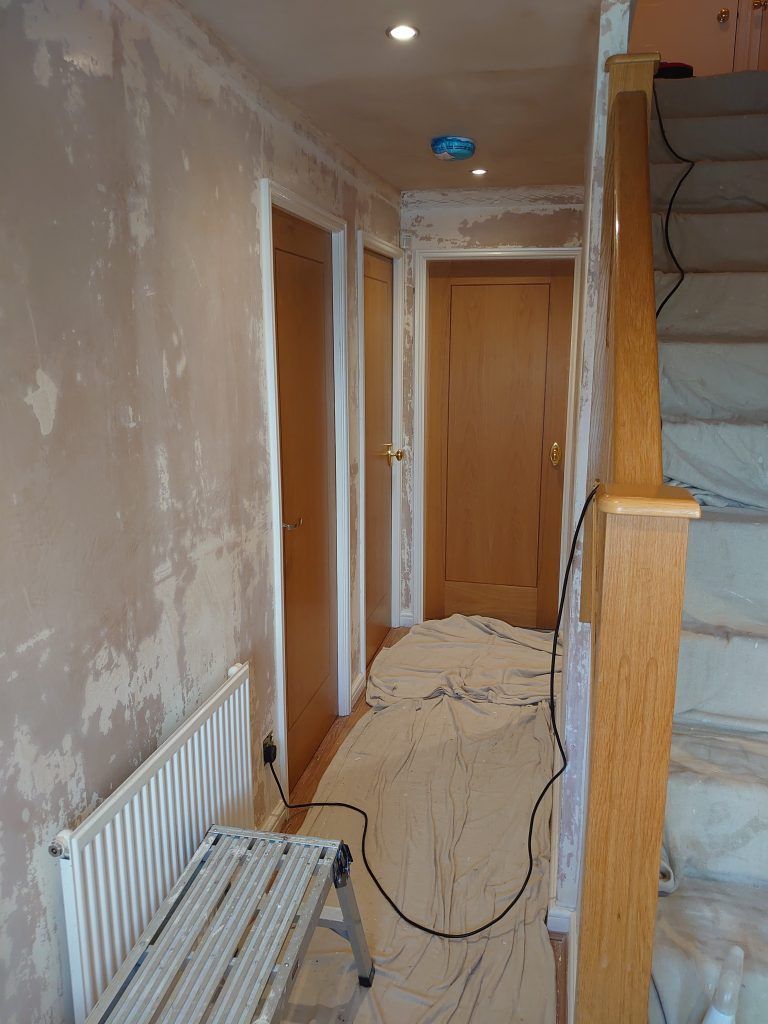 A hallway under renovation with bare plaster walls, wood doors, a radiator, and a step stool on a protective floor sheet.