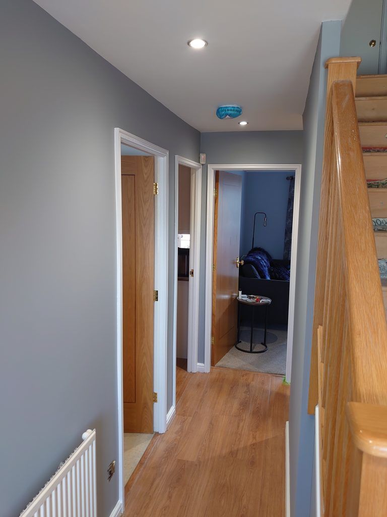 A hallway with grey walls and wood-look flooring, leading to doorways and a wooden staircase.
