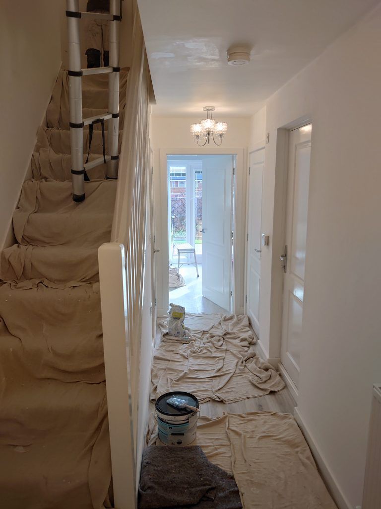 A hallway under renovation with drop cloths covering the floor and stairs, a paint can, and a telescopic ladder.