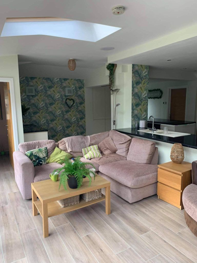 A living room with a pink sectional sofa, wooden coffee table, a green plant, and botanical wallpaper in a bright space.