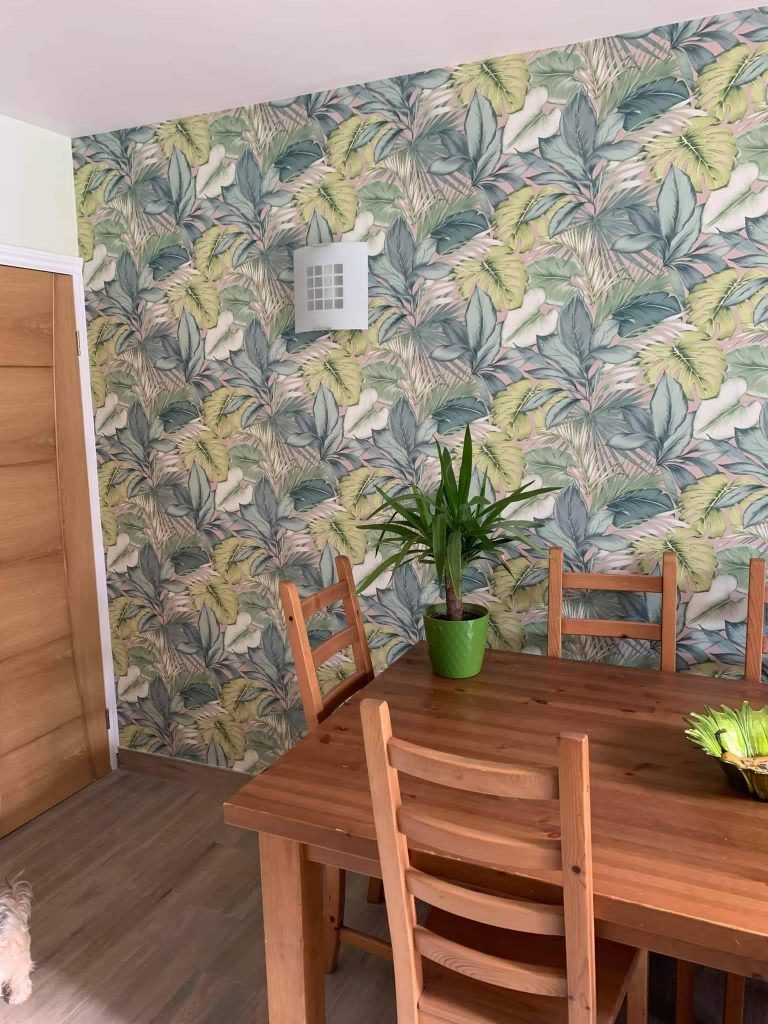 A wooden dining table and chairs set against a wall with tropical leaf-patterned wallpaper and a green potted plant.