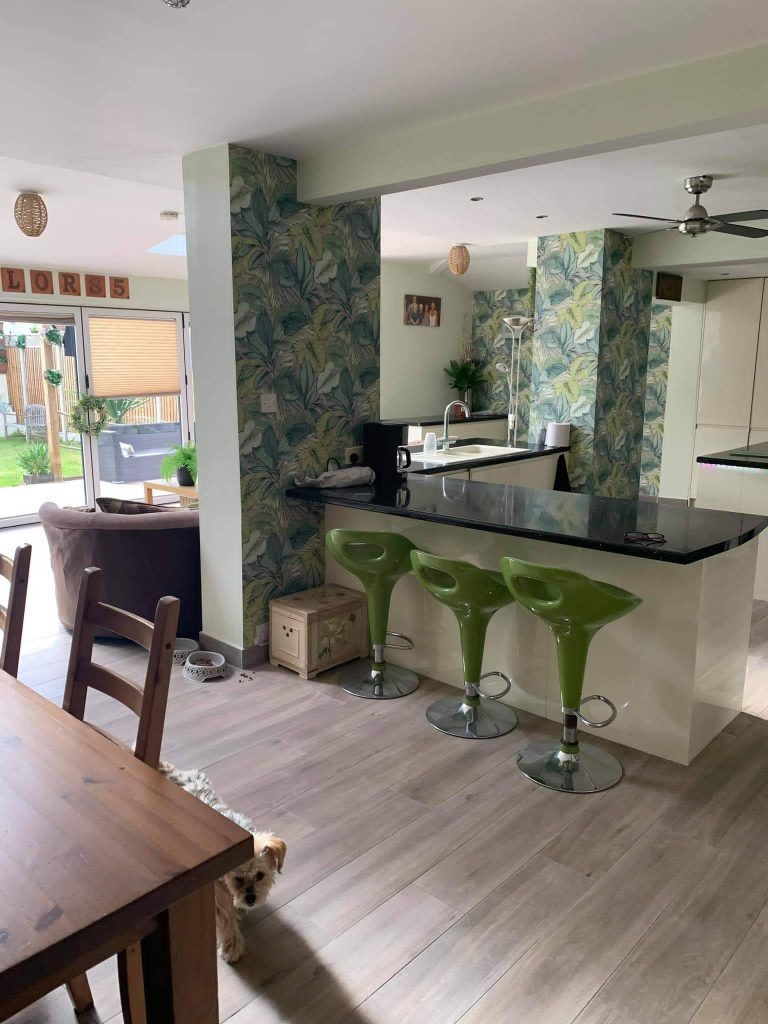A kitchen with a black breakfast bar, three lime green stools, and botanical wallpaper, seen from a dining area with a dog.