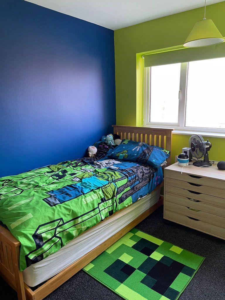 A child's bedroom featuring a blue wall, green wall, Minecraft-themed bedding, a wooden bed frame, and a pixelated rug.