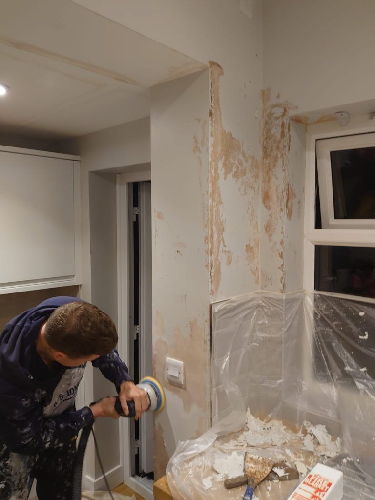 A person wearing a dark hoodie uses a sanding tool on a partially stripped, light-colored kitchen wall.
