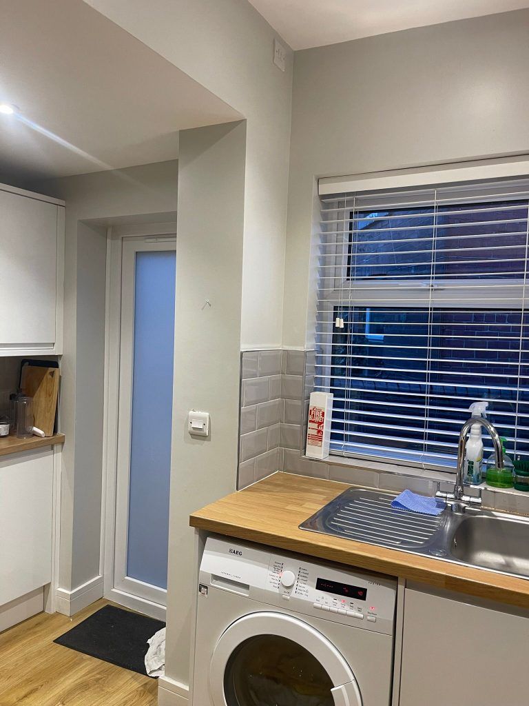 A bright kitchen with white cabinets, a light wood countertop, a washing machine, a window with blinds, and a frosted door.