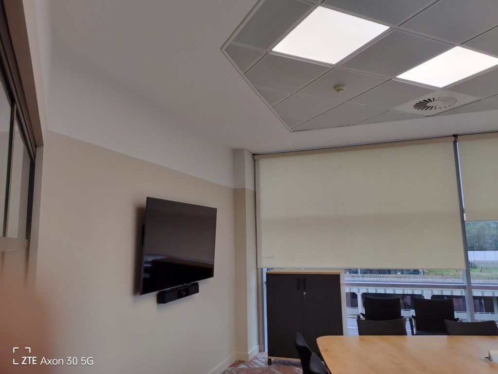 A conference room with a wall-mounted TV, beige window blinds, a wooden table, and chairs under drop ceiling lighting.