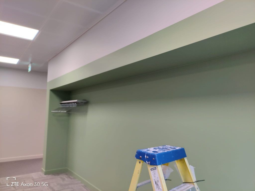 A ladder stands in an office space with a recessed green wall featuring a mounted shelf.