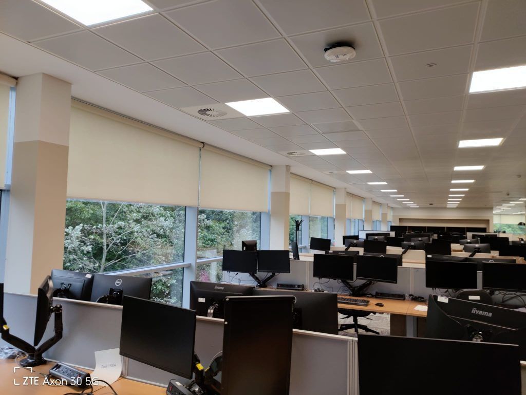 Empty office with rows of cubicles, monitors, and large windows with closed shades.