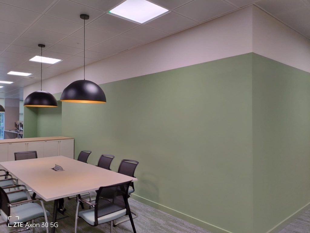 A modern office meeting room with a large table, black chairs, two hanging dome lights, and sage green walls.