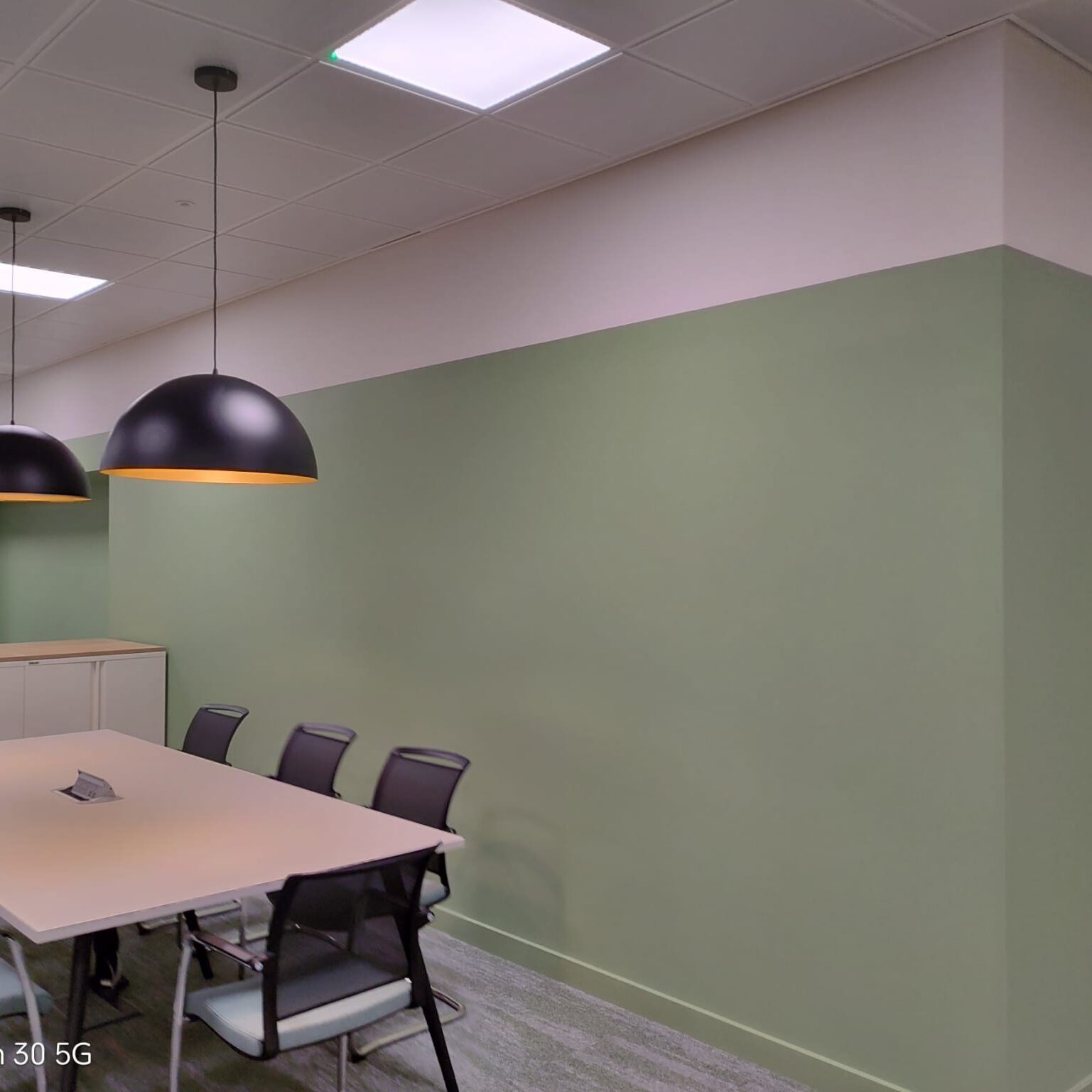 Modern office meeting room with a light table, black office chairs, two pendant lights, and a sage green accent wall.