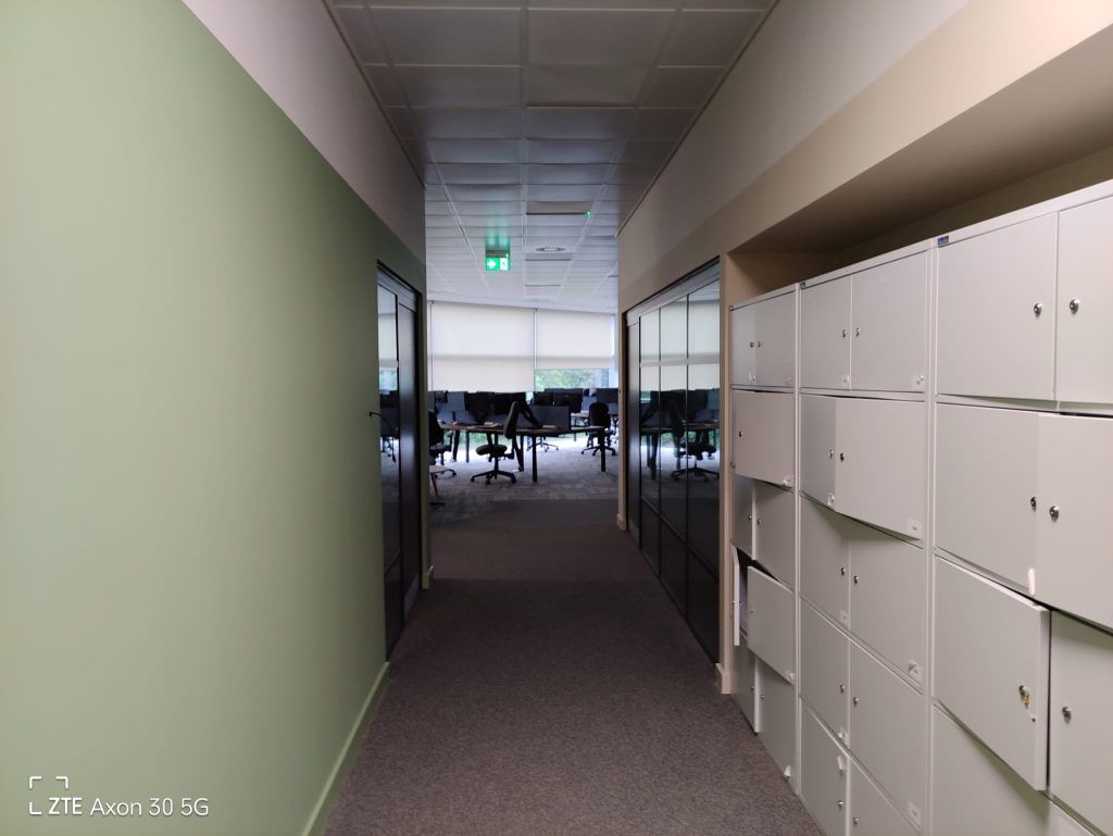 A carpeted office hallway with a sage green wall on the left, white lockers on the right, and an open workspace beyond.