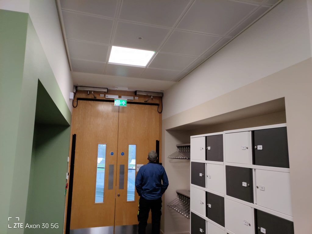 A person stands in a hallway facing a pair of wooden double doors with an exit sign, next to a wall of storage lockers.