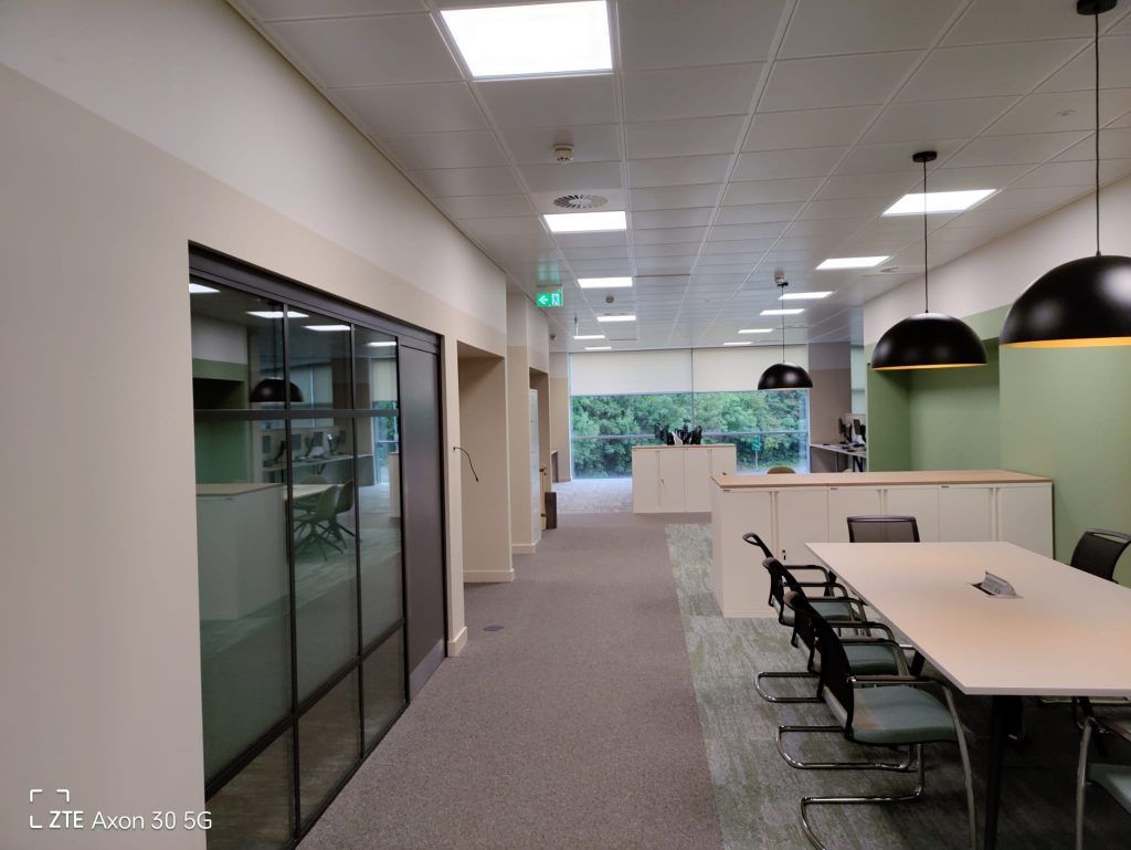 A modern office hallway with a glass-walled conference room on the left and a white table with chairs on the right.