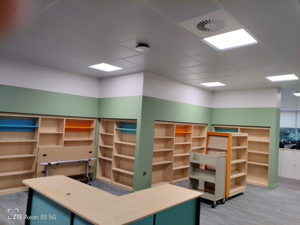 A modern library space with light wood bookshelves, sage green walls, and an L-shaped service desk in the foreground.
